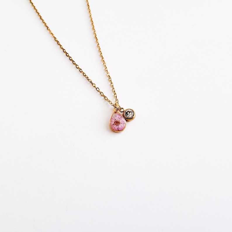 Op de afbeelding: Een delicate goudkleurige ketting met een druppelvormige hanger met een roze bloem. Een kleine, ronde, heldere edelsteen hangt naast de hanger. De ketting staat op een witte achtergrond.
