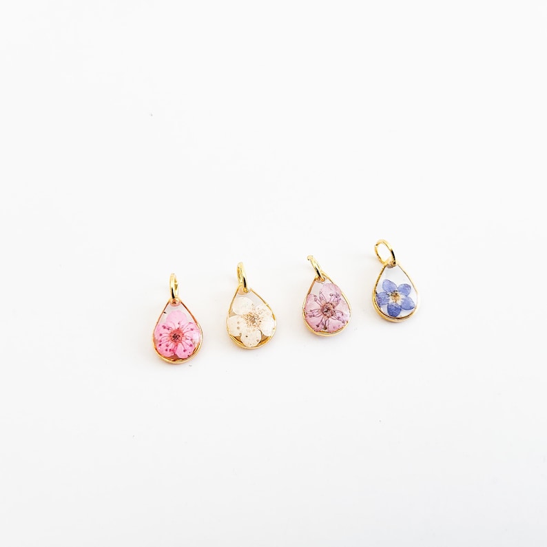 Op de afbeelding: Vier druppelvormige hangers met goudkleurige frames, elk met een andere geperste bloem. De bloemen zijn roze, wit, roze en blauw. De hangers worden op een witte achtergrond weergegeven.