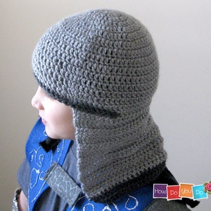 May include: A gray crocheted hat with a long flap that covers the neck and ears. The hat is made with a simple stitch pattern and has a black trim around the edge.