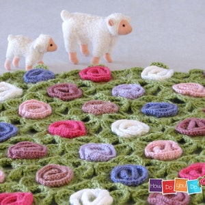 Puede incluir: Una manta de ganchillo verde con un diseño floral que presenta múltiples rosas de ganchillo de color pastel. Dos corderos de peluche blancos están en el fondo.
