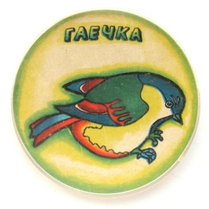 May include: A round, yellow, green, blue, and red toy with a bird illustration and the word "ГАЕЧКА" printed on it.