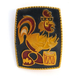Puede incluir: Un pin de esmalte dorado con un gallo de dibujos animados tocando un tambor. El gallo es rojo, amarillo y verde, y el tambor es rojo y blanco con puntos negros.