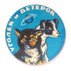 May include: A circular sticker featuring a blue background with a graphic illustration of two dogs. The text "УГОЛЕК И ВЕТЕРОК" is printed in white along the upper edge, and a small blue planet with white orbital rings is visible above the dogs.