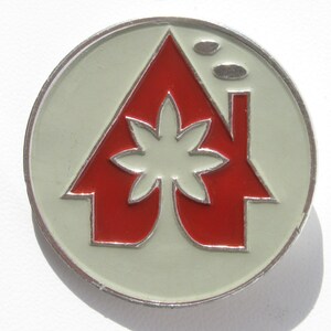 May include: A silver pin with a red house with a white flower in the center. The house has a chimney with two smoke puffs above it.