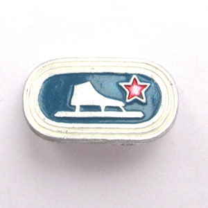 May include: A small, oval-shaped pin with a white border and a blue interior. The pin features a white ice skate and a red star. The ice skate is on a blue background.