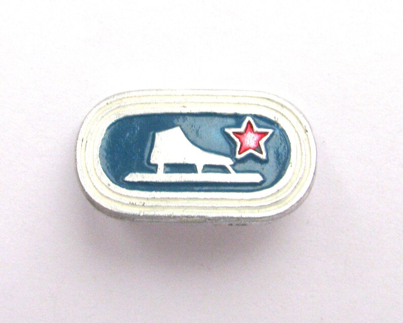 May include: A small, oval-shaped pin with a white border and a blue interior. The pin features a white ice skate and a red star. The ice skate is on a blue background.