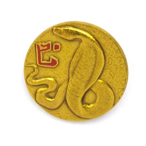 Puede incluir: Un pin de metal dorado con una imagen en relieve de una cobra. La serpiente está enroscada y tiene un carácter chino rojo y dorado en su espalda. El pin es circular y tiene un fondo texturizado.