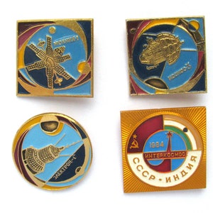 May include: Four gold-colored pins with colorful designs depicting space exploration. The pins feature a satellite, a space station, a rocket, and a logo with the text "1984 INTERKOSMOS СССР - ИНДИЯ".