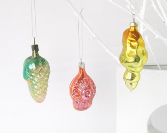 May include: Three vintage glass Christmas ornaments hanging from white branches. The ornaments are a green and white grape cluster, a pink and white swirl design, and a yellow and gold teardrop shape.