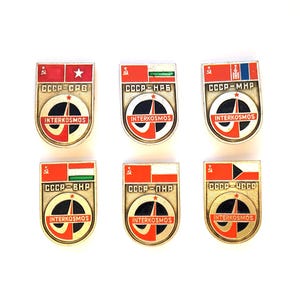 May include: A set of six metal Interkosmos pins featuring the Soviet hammer and sickle, various national flags, and the text CCCP combined with country abbreviations. Each shield-shaped badge displays a central circular logo with a red star.