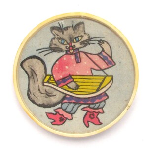 以下が含まれることがあります： 丸い木製の玩具で、ピンクと白のドレスを着た灰色の猫が黄色の楽器を演奏している絵が描かれています。猫は青い目と赤い口をしています。