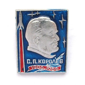 Op de afbeelding: Een rechthoekige metalen speld met een zilveren profielportret van S.P. Korolev tegen een blauwe emaille achtergrond met zilveren sterren. Een zilveren raket stijgt links op en een rode banner onderaan toont de tekst S.P. KOROLEV 1906-1966.