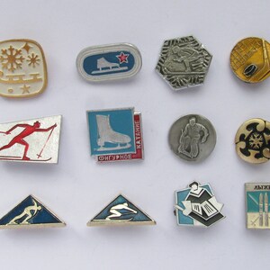 May include: A collection of vintage enamel pins featuring winter sports themes. Designs include ice skates, skiers, snowflakes, and hockey. The pins are in various shapes, including squares, ovals, and triangles, with a mix of gold, silver, red, blue, and white colors.