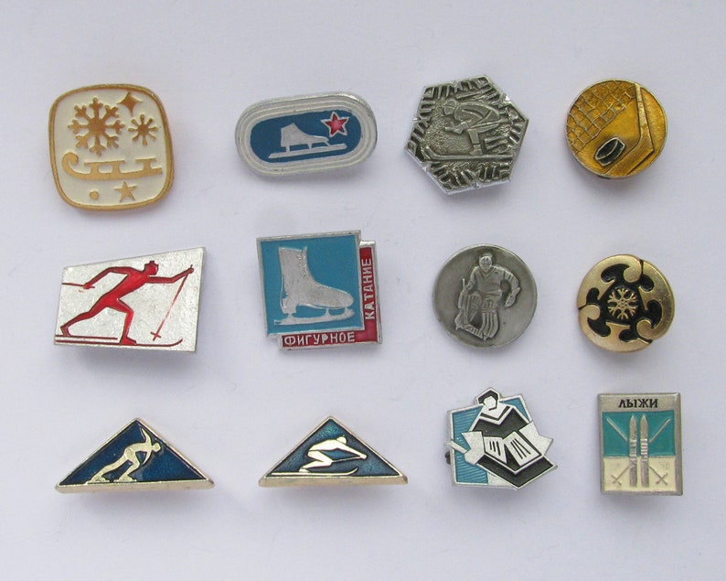 May include: A collection of vintage enamel pins featuring winter sports themes. Designs include ice skates, skiers, snowflakes, and hockey. The pins are in various shapes, including squares, ovals, and triangles, with a mix of gold, silver, red, blue, and white colors.