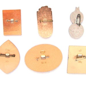 May include: A collection of six metal pins shown from the reverse side, each featuring a horizontal pin-back closure. The pins vary in shape, including rectangular, oval, and leaf-like forms, with some displaying stamped text and numerical markings.