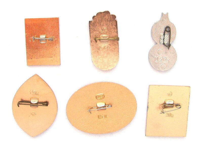 May include: A collection of six metal pins shown from the reverse side, each featuring a horizontal pin-back closure. The pins vary in shape, including rectangular, oval, and leaf-like forms, with some displaying stamped text and numerical markings.