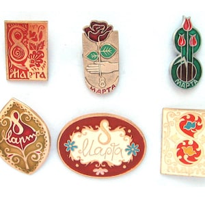 May include: A collection of six metal enamel pins featuring various designs with the text 8 Marta. The pins include rectangular, oval, and leaf shapes in red, gold, green, and white colors, each commemorating the date with floral and graphic motifs.