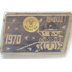 May include: A rectangular pin with a clear plastic border featuring a dark blue background with a white graphic of the Lunokhod 1 lunar rover. The text 1970, LUNOKHOD 1, and a circular emblem with the letters CCCP and the year 1958 are visible.