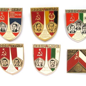 May include: Six vintage enamel pins featuring Soviet space missions. The pins are shaped like shields and feature red, white, and blue colors. The pins depict astronauts and the text "CCCP" and "COIO3".