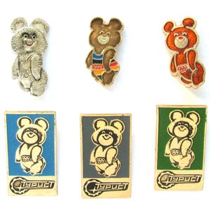 May include: Six enamel pins featuring a cartoon bear wearing a swimsuit. The bear is in different colors, including silver, orange, and brown. The pins are rectangular and have the word "турист" written on them.