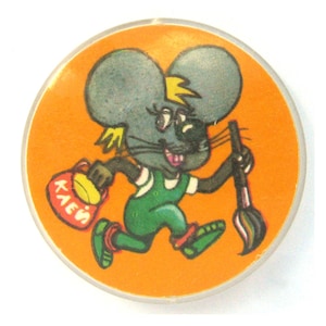Puede incluir: Un botón redondo naranja con un dibujo animado de un ratón. El ratón, con un mono verde, sostiene un pincel y una bolsa con texto. El botón tiene un borde de plástico transparente.