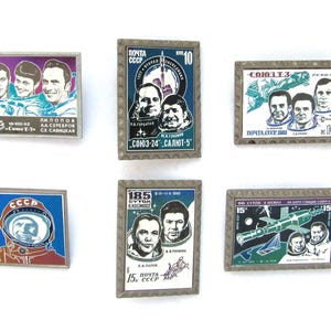 May include: A collection of six vintage Soviet space-themed pins. Each pin features images of cosmonauts, spacecraft, and text in Russian. The pins are rectangular and square, with silver-colored borders and colorful enamel designs.