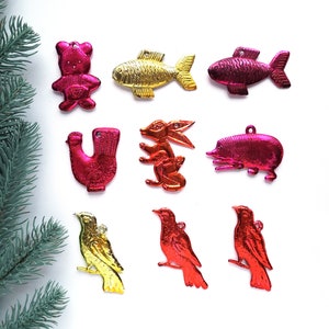 Könnte beinhalten: Eine Sammlung bunter, metallischer Weihnachtsschmucke in verschiedenen Formen, darunter ein Bär, Fische und Vögel. Die Ornamente sind in Gold- und Rottönen gehalten und haben eine glänzende, reflektierende Oberfläche. Ein Zweig eines Nadelbaums ist in der Ecke.