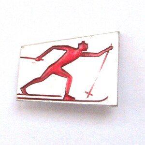 May include: A rectangular pin featuring a red skier in motion against a white background. The skier is depicted with skis and poles, suggesting a winter sports theme. The pin has a simple, vintage design.