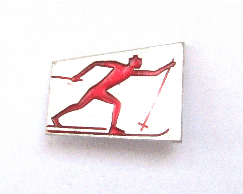 May include: A rectangular pin featuring a red skier in motion against a white background. The skier is depicted with skis and poles, suggesting a winter sports theme. The pin has a simple, vintage design.