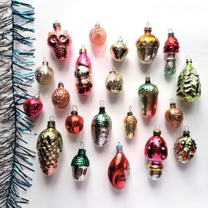 May include: A collection of vintage Christmas ornaments in various shapes and colors, including a skull, mushroom, acorn, and pinecone. The ornaments are primarily pink, red, gold, and green, with a reflective, glass-like finish. Tinsel is visible in the background.