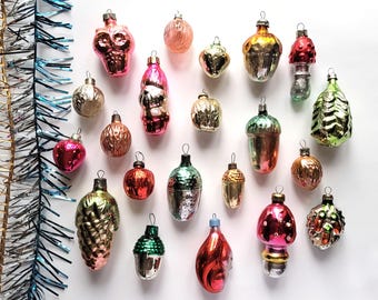 May include: A collection of vintage Christmas ornaments in various shapes and colors, including a skull, mushroom, acorn, and pinecone. The ornaments are primarily pink, red, gold, and green, with a reflective, glass-like finish. Tinsel is visible in the background.