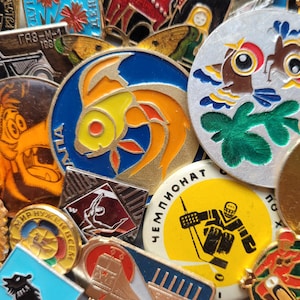 Può includere: Una collezione di spille vintage in metallo smaltato con vari disegni, tra cui un pesce dorato su sfondo blu, un giocatore di hockey su sfondo giallo, un'auto, un lupo e forme astratte con testo in cirillico su diversi pezzi.