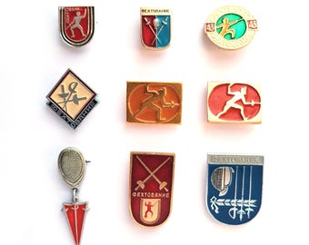 May include: A collection of nine metal pins featuring fencing themes, including various shield, diamond, and rectangular shapes. The pins display enamel colors of red, blue, orange, and green, with metallic gold and silver accents depicting fencers.