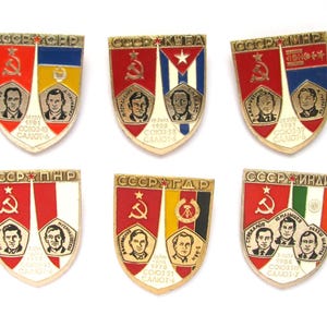 May include: Six vintage Soviet-era pins with shield shapes. Each pin features a red star, hammer and sickle, and portraits of cosmonauts. The pins have flags of different countries and Cyrillic text.