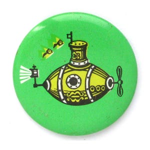 Peut inclure: Un bouton rond vert avec une illustration de sous-marin jaune et des accents noirs et blancs. Le sous-marin a une hélice, un périscope et un hublot. Deux poissons jaunes nagent au-dessus du sous-marin.