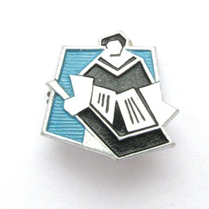 May include: A vintage enamel pin featuring a stylized illustration of a person in a graduation gown reading a book. The pin has a black, white, and light blue color scheme with a silver border. The pin is likely a collectible item or a souvenir.