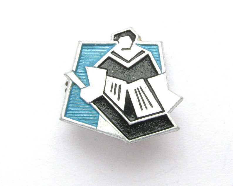 May include: A vintage enamel pin featuring a stylized illustration of a person in a graduation gown reading a book. The pin has a black, white, and light blue color scheme with a silver border. The pin is likely a collectible item or a souvenir.