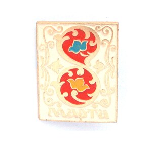 May include: A rectangular metal pin featuring a cream-colored background with gold-toned filigree borders. The center displays two stacked circular red motifs containing a blue and a yellow bird-like shape. The Cyrillic text "МАРТА" is at the bottom.