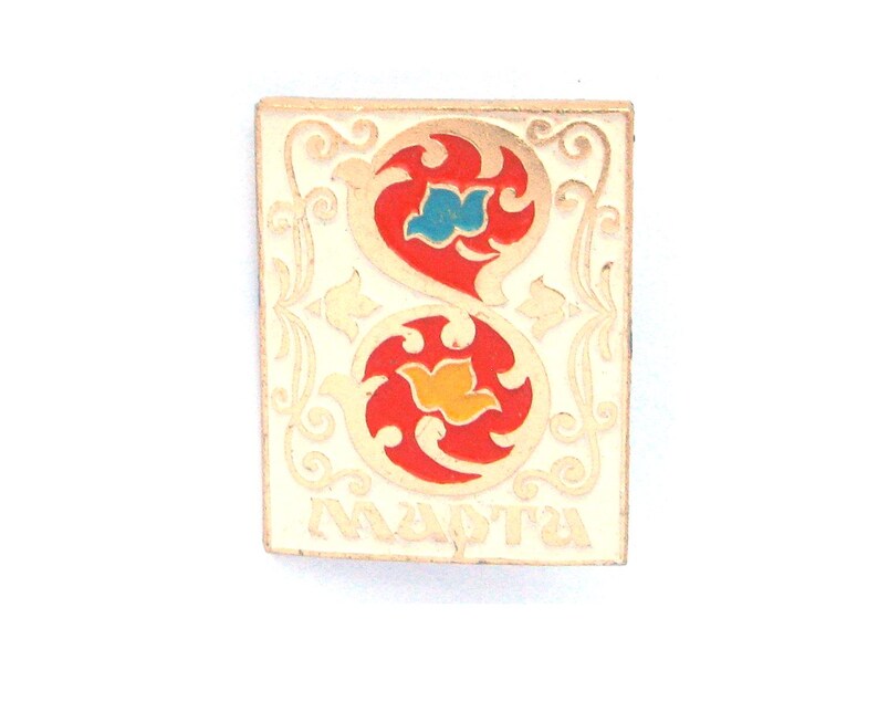 May include: A rectangular metal pin featuring a cream-colored background with gold-toned filigree borders. The center displays two stacked circular red motifs containing a blue and a yellow bird-like shape. The Cyrillic text "МАРТА" is at the bottom.