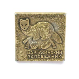 Puede incluir: Pin metálico cuadrado, dorado, con un animal estilizado y texto cirílico. El pin tiene un fondo texturizado y un diseño detallado, probablemente un recuerdo o artículo de colección vintage.