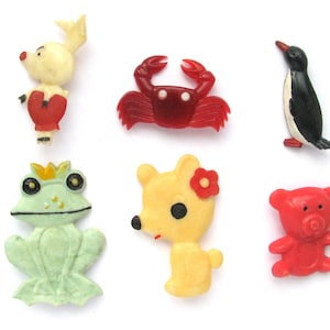 May include: A collection of six colorful, small animal-shaped figurines. The figurines include a white rabbit with red accents, a red crab, a black and white penguin, a green frog with a crown, a yellow bear with a red flower, and a red teddy bear.
