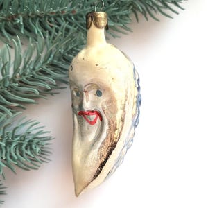 May include: Vintage Christmas ornament shaped like a crescent moon with a face. The ornament has a cream-colored base with silver accents, blue details, and a red mouth. A gold-colored metal cap is at the top.