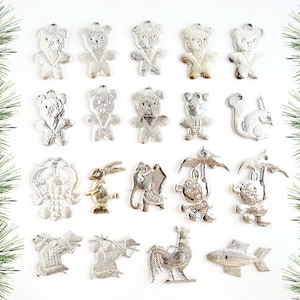 May include: A collection of silver-colored metal ornaments in various animal shapes, including bears, a squirrel, a rabbit, monkeys with umbrellas, and a fish. The ornaments are arranged on a white surface, with green pine branches framing the image.