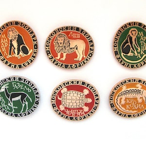 以下が含まれることがあります： 黒い縁取りのある丸い金色のピンが6つ。それぞれのピンには、ライオン、サル、ガゼル、カメ、ワニ、マンドリルなど、アフリカの動物が描かれています。ピンにはすべて、キリル文字で「Moskovskiy Zoopark」と「Fauna Afriki」と書かれています。