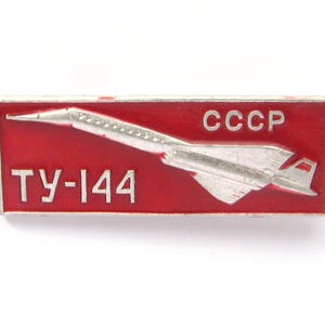 Pode incluir: Um alfinete vermelho e prateado com um avião branco e o texto "CCCP" e "TY-144" nele.