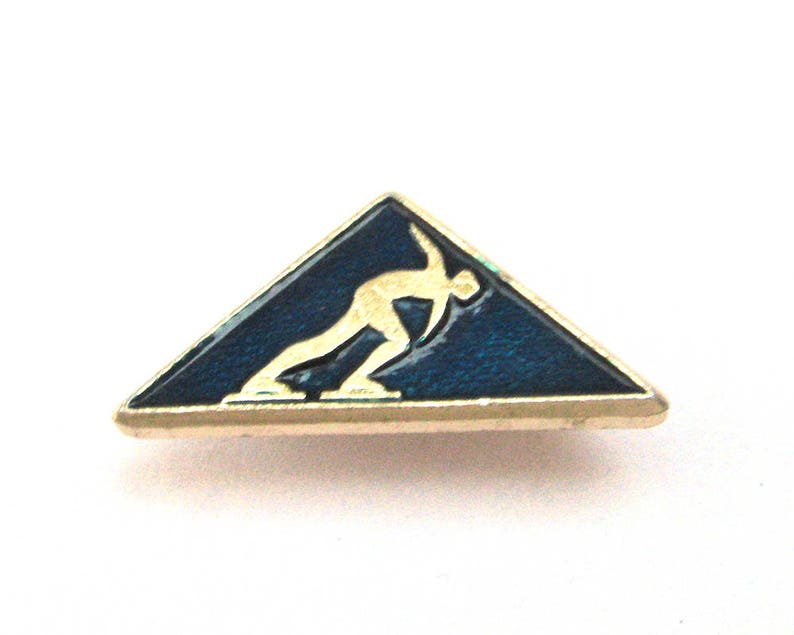 May include: A vintage pin in the shape of a triangle, featuring a gold-colored border. The pin has a dark blue enamel background with a gold-colored figure of a speed skater. A collectible sports-themed accessory.