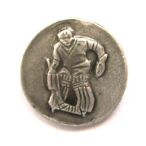 May include: A silver-toned metal button featuring a relief of a cricket player in action. The player is depicted in a batting stance, wearing protective gear. The button is round and has a slightly aged appearance.