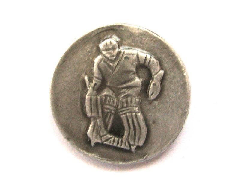 May include: A silver-toned metal button featuring a relief of a cricket player in action. The player is depicted in a batting stance, wearing protective gear. The button is round and has a slightly aged appearance.