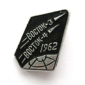 Może przedstawiać: Metalowa odznaka w kolorze czarnym i srebrnym z napisem "Wostok-3" i "Wostok-4/1962". Odznaka przedstawia rakietę startującą w kosmos.