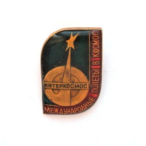 Puede incluir: Pin espacial vintage de Interkosmos con un diseño dorado y verde oscuro. El pin presenta una estrella, un cohete y el texto cirílico "Интеркосмос" y "МЕЖДУНАРОДНЫЕ ПОЛЕТЫ В КОСМОС". El pin tiene un borde dorado.